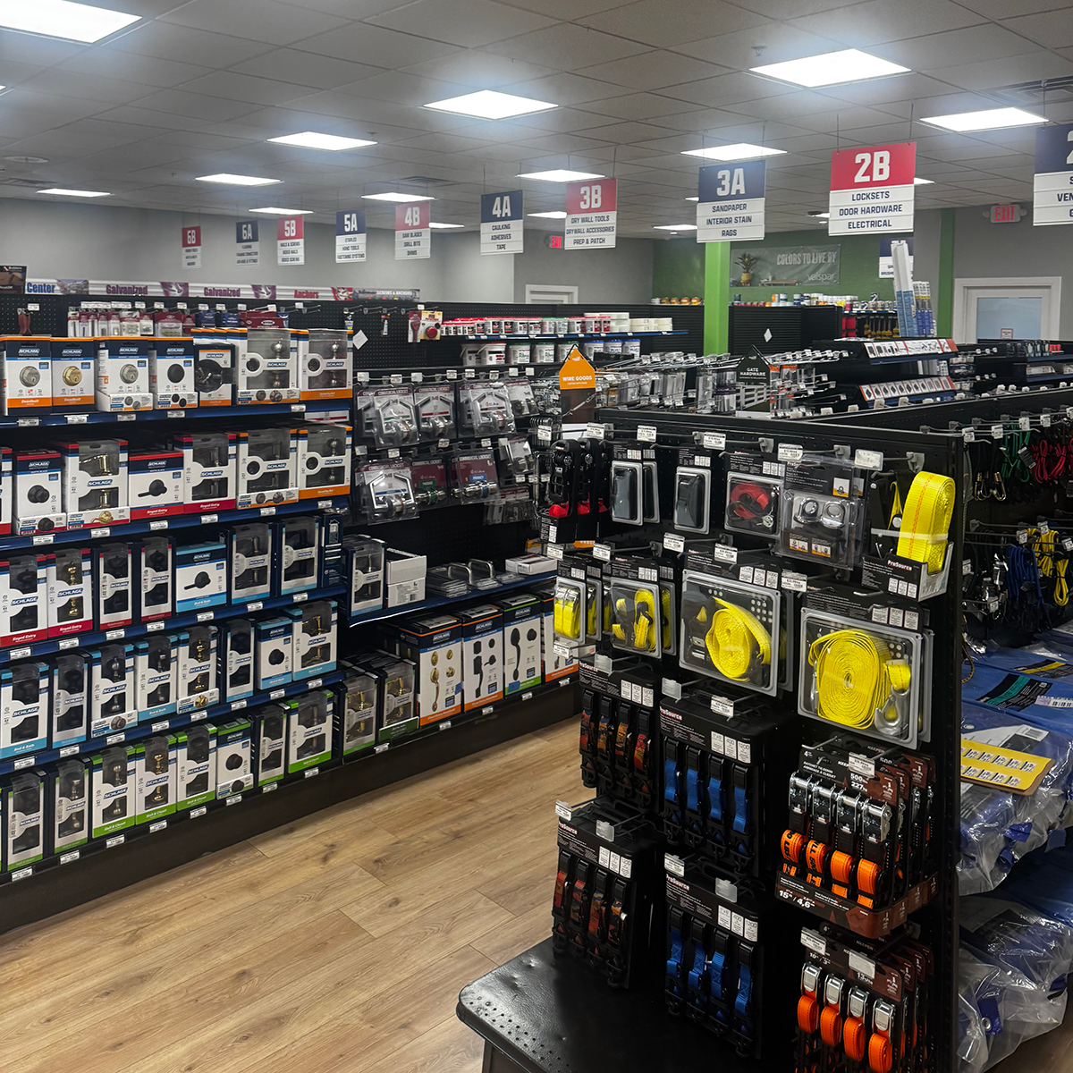 Wide interior view of a Builders FirstSource store showing rows of locksets, door hardware, electrical supplies and colorful tie-down straps. Shelves at left hold boxed Schlage hardware, while a black center display holds packaged straps and other accessories. Overhead signs identify sections for locksets, door hardware, electrical, sandpaper, interior stain, rags, dry wall tools and accessories