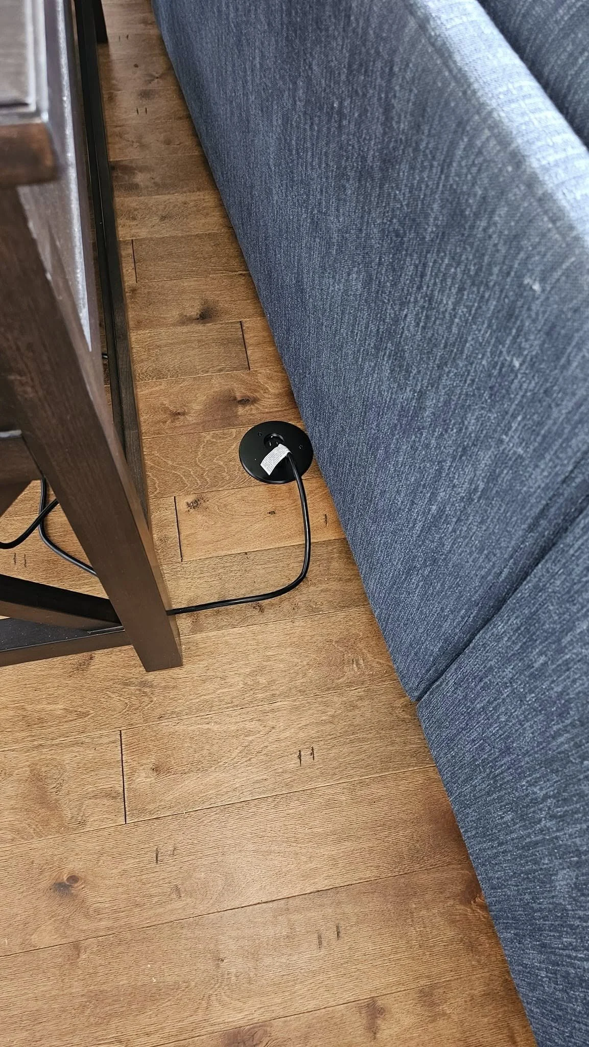 A black power cord runs from a floor outlet, nestled against the leg of a dark wood table and the side of a blue upholstered sofa. The outlet has a white label attached. The floor is made of light brown wood planks with visible grain and knots.