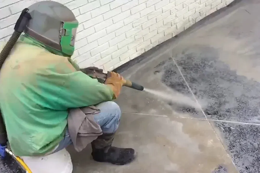 Worker performs surface cleaning on concrete floor using blasting equipment, removing dirt, coatings, and stains, ensuring proper preparation for sealing, coating, or resurfacing in residential or commercial settings.