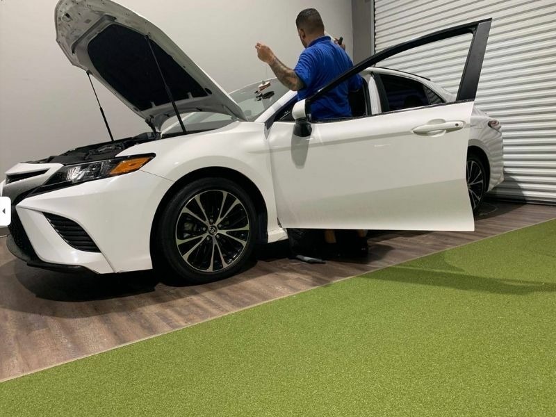 Technician prepping a sedan for a windshield replacement, hood up to protect the cowl and wipers during the job.