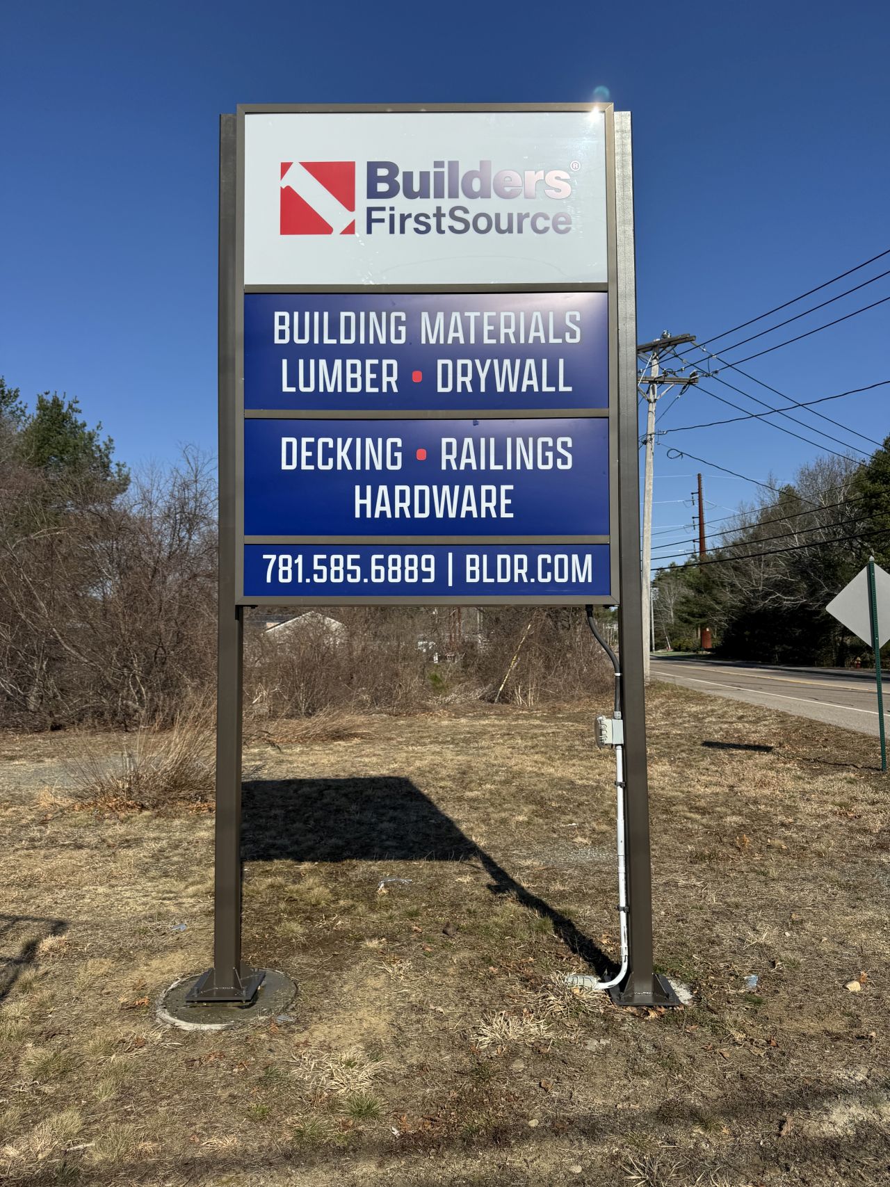 Freestanding Builders FirstSource roadside sign listing building materials, lumber, drywall, decking, railings, and hardware, positioned along a roadway near the lumber supply location.
