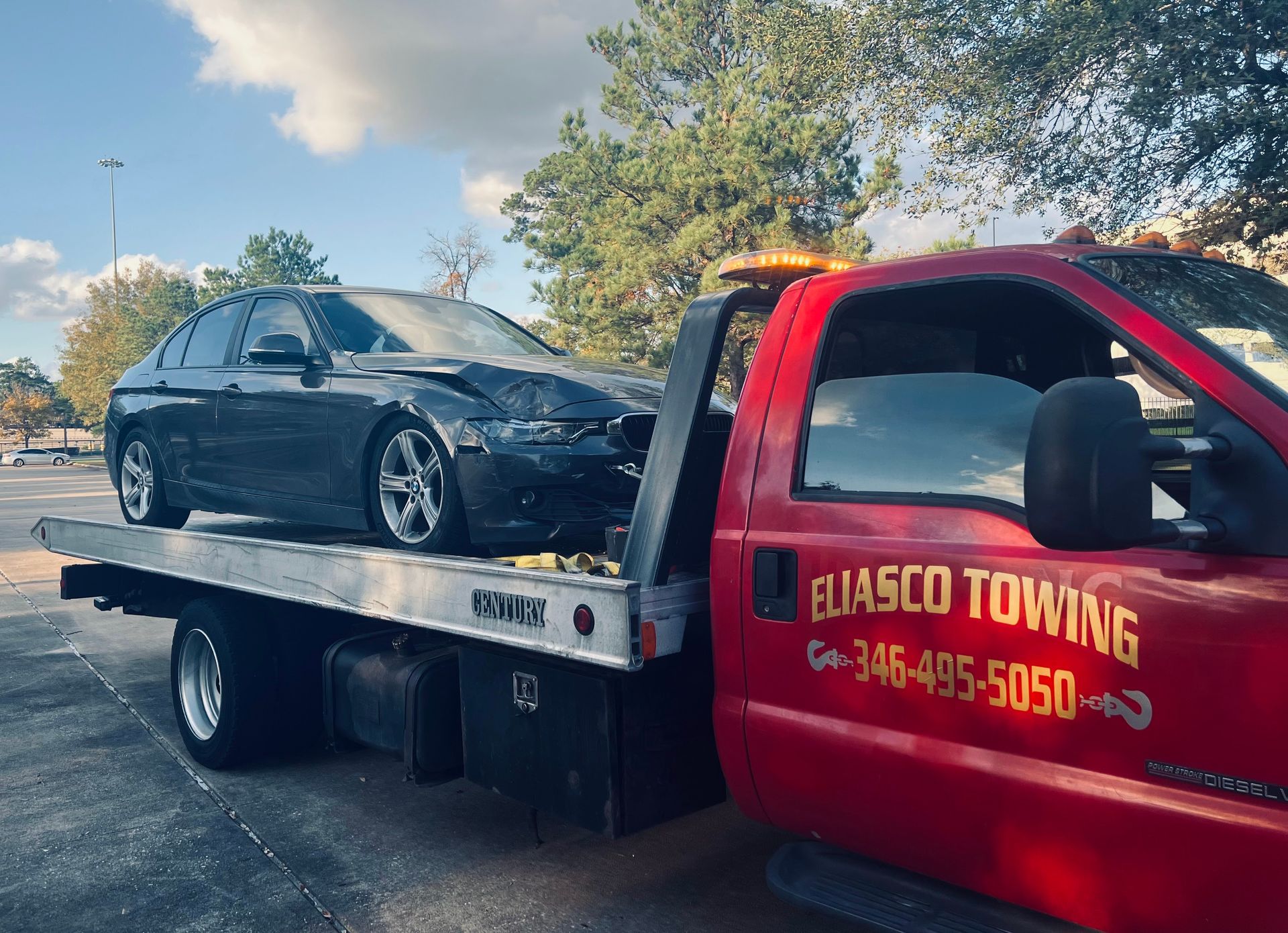 An Eliasco Towing flatbed truck transports a gray BMW sedan with visible front-end collision damage, loaded and secured on the aluminum bed near trees and a parking lot.