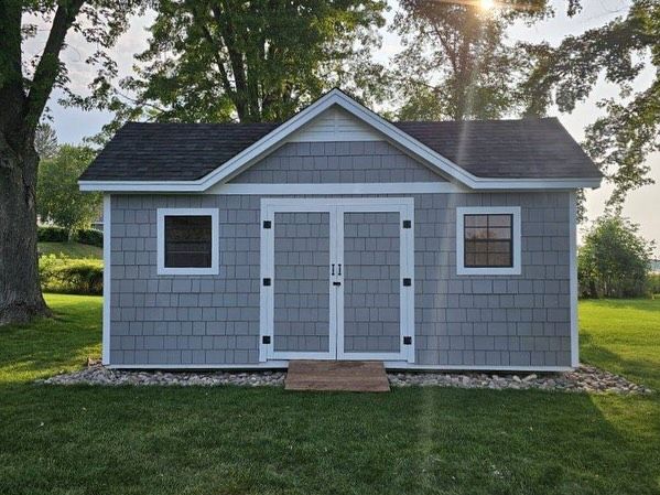 A stylish, pre-built storage shed featuring gray scallop or shake-style siding, white trim around the windows and double doors, and a black shingled roof. It sits on a grassy yard over a small bed of gravel.