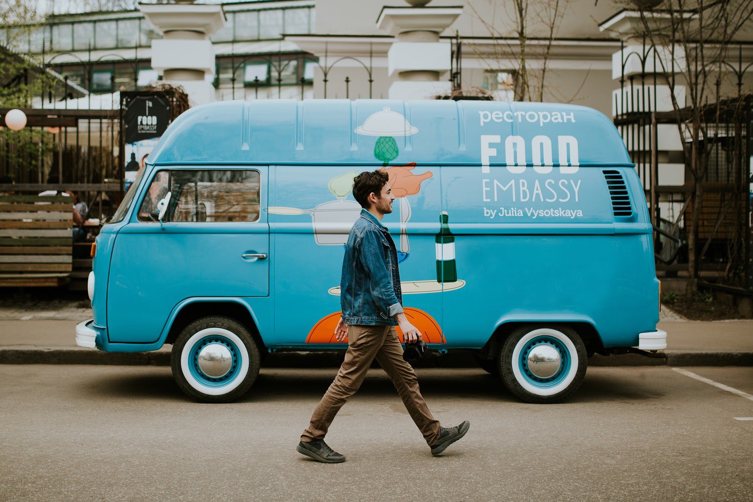 A man walks past a bright blue vintage Volkswagen van with "FOOD EMBASSY by Julia Vysotskaya" written on its side. The van is decorated with illustrations of kitchenware and food items. The man is wearing a denim jacket and brown pants, and is walking from left to right, looking towards the van. The background shows a building and some trees, suggesting an urban or suburban outdoor setting.