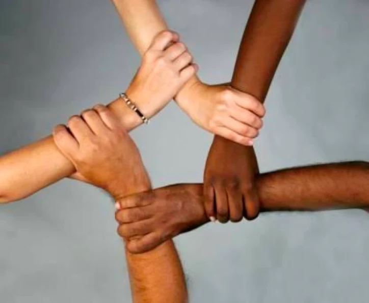 A close-up shot shows four arms and hands interlocked in a circle, symbolizing unity and cooperation. The hands are of varying skin tones, suggesting diversity, and one hand wears a bracelet.
