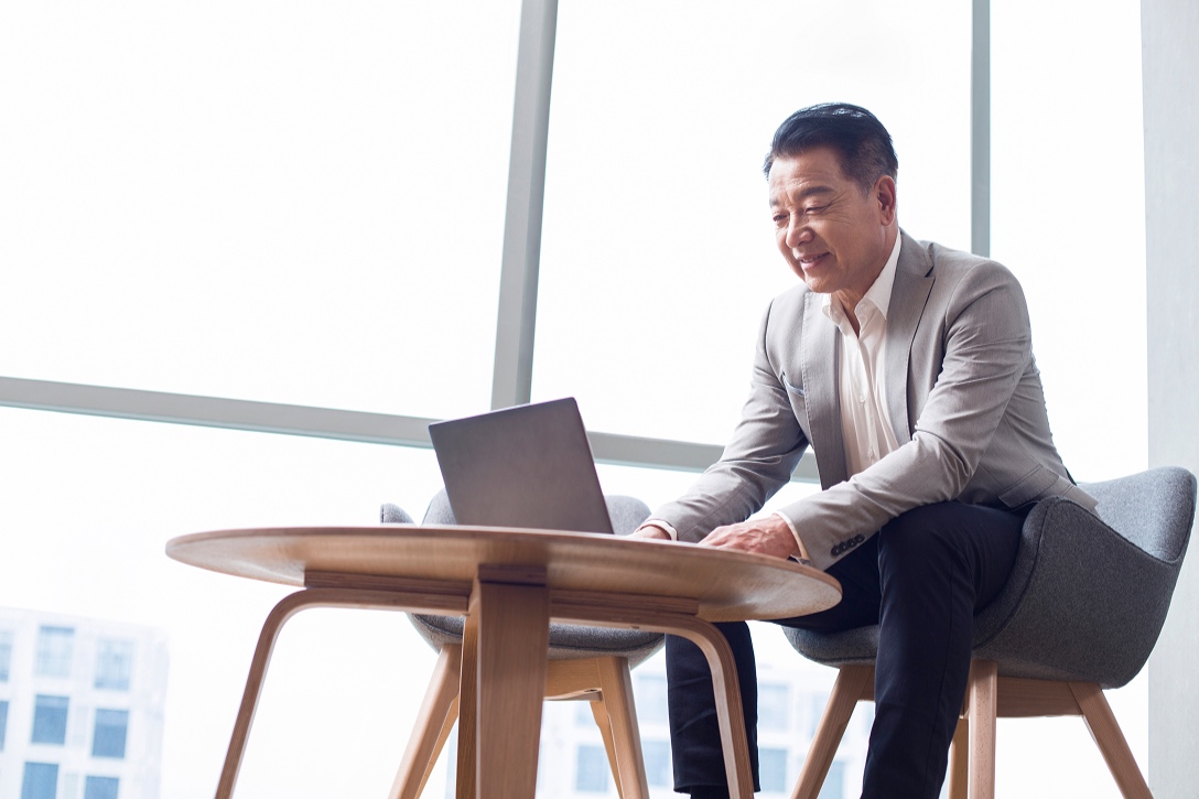 A man in a gray suit sits on a gray chair, smiling and using a laptop on a wooden table. Bright, modern office with large windows in the background.