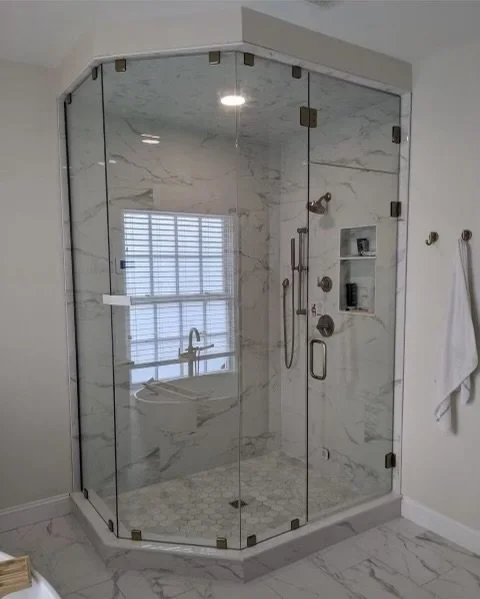 A modern bathroom with a large, multi-panel glass shower enclosure with a white marble tile interior. Inside the shower, a rainfall shower head, handheld shower, and a built-in shelf are visible. The shower floor is tiled with small hexagonal white tiles. Adjacent to the shower, a white bathtub is partially visible behind the glass, and a window with blinds is to the left. The bathroom floor is tiled with large, white marble-patterned tiles. Two hooks with white towels hang on the wall to the right of the shower.
