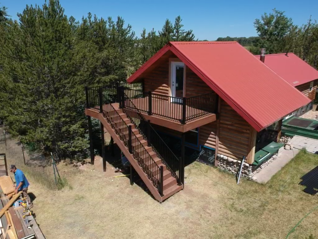This aerial shot captures a custom project by DeckPro LLC designed to perfectly complement a rustic log cabin. The second-story composite deck features durable materials, sleek black metal railing, and a sturdy staircase connecting the upper level to the yard below.
