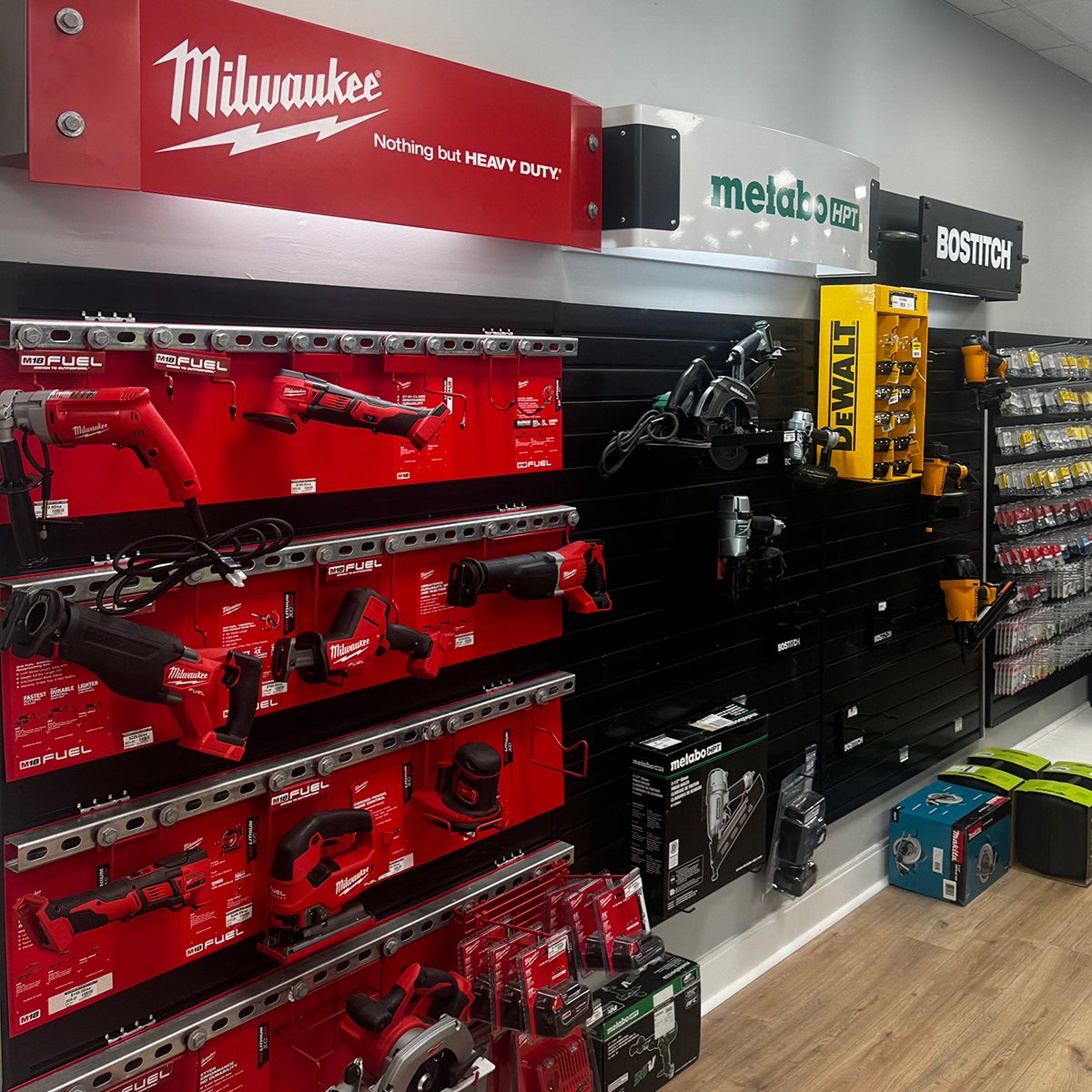 Builders FirstSource showroom display featuring red Milwaukee tools beneath a “Nothing but HEAVY DUTY” sign, alongside Metabo HPT and Bostitch power tools. Corded saws, drills, rotary tools and other equipment are mounted on black wall panels with labeled product packaging. Yellow DEWALT products and rows of small accessories appear along the right side.
