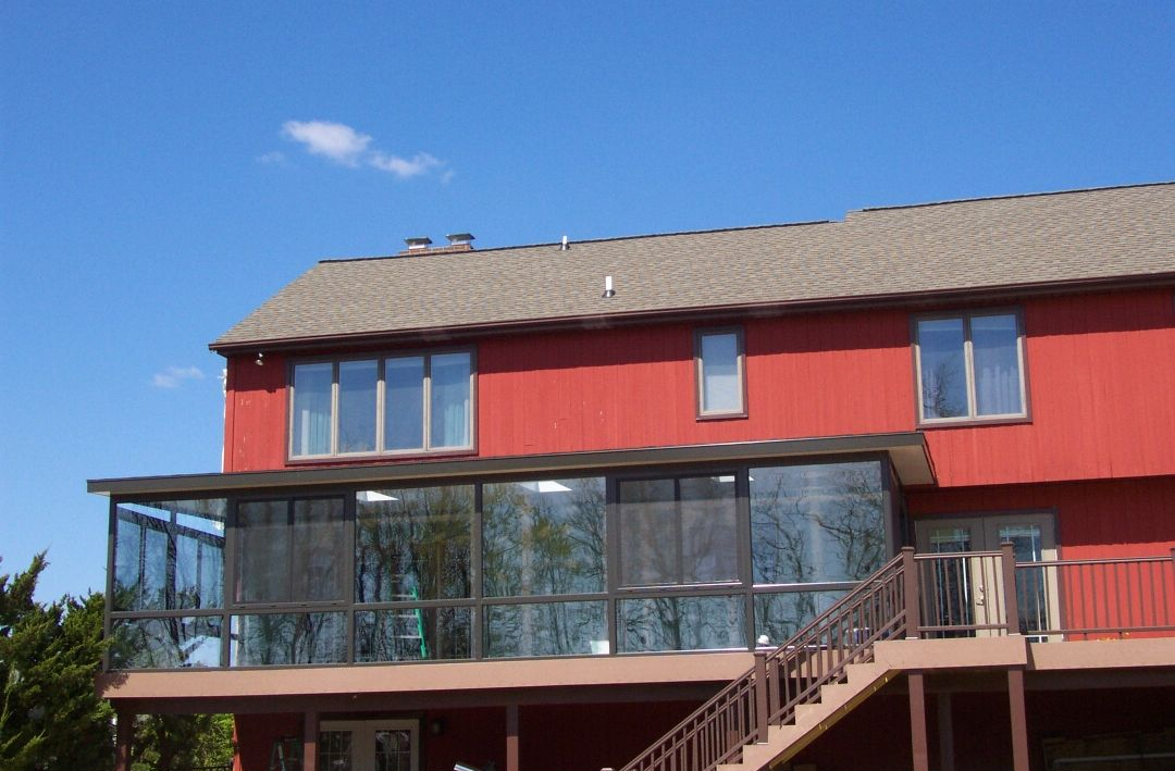 Large elevated screened-in porch attached to a red two-story home, featuring dark framing, expansive mesh panels, and a sloped roofline. The structure extends outdoor living space, offering panoramic backyard views while providing protection from insects and weather conditions.