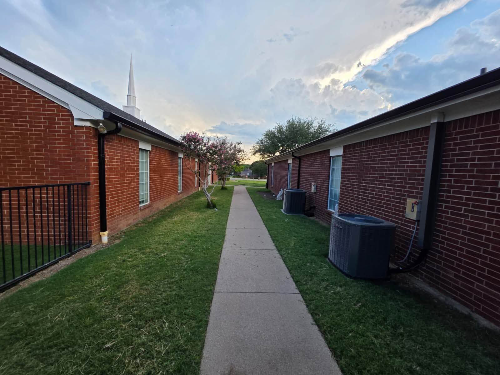 Commercial property exterior featuring brick buildings, dark shingle roofs, central concrete walkway 