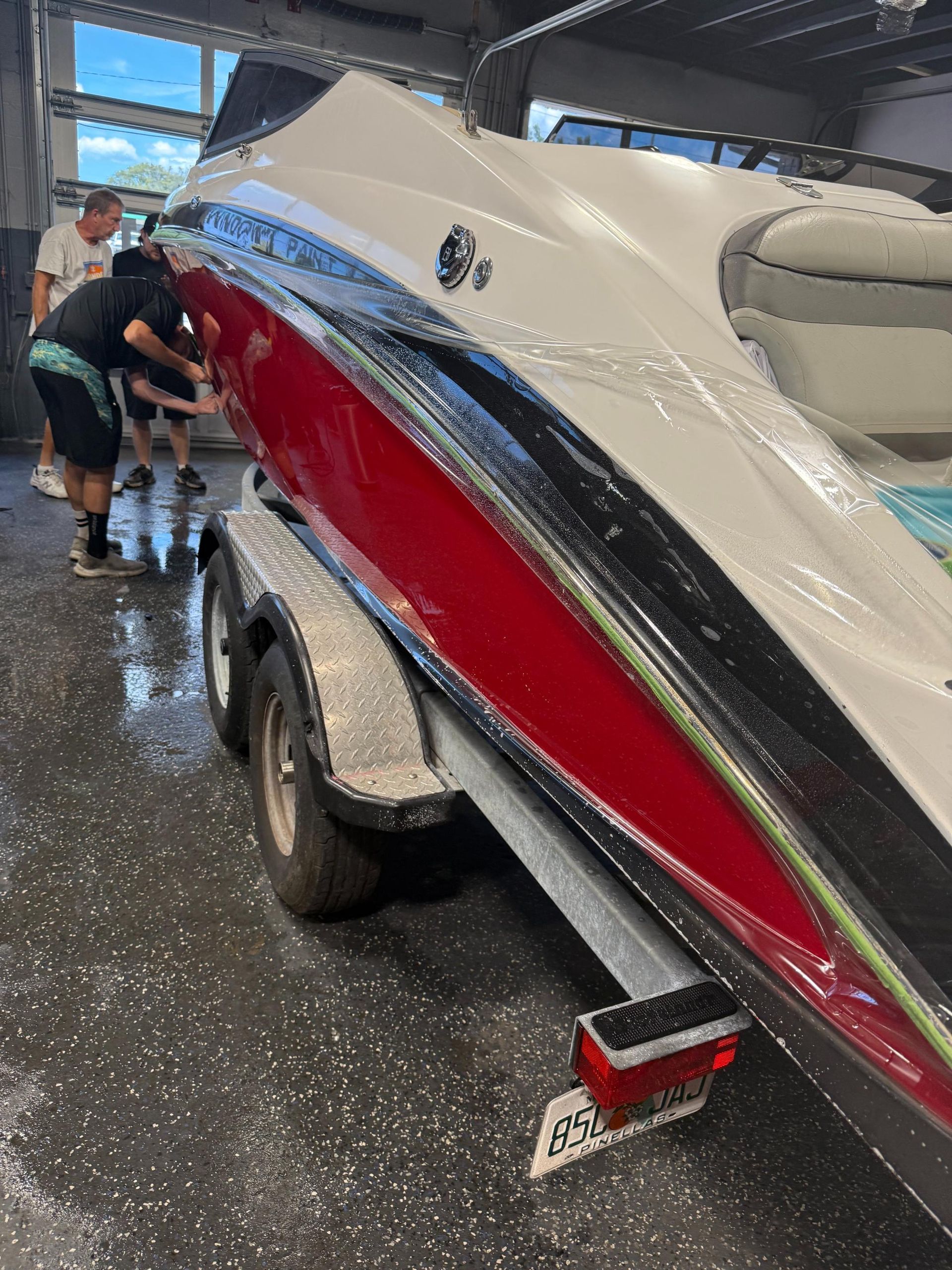 Several technicians are actively working on the exterior of a boat hull, which is white and red, while it rests on a trailer inside a service bay. The boat's surface is partially covered in protective plastic film.