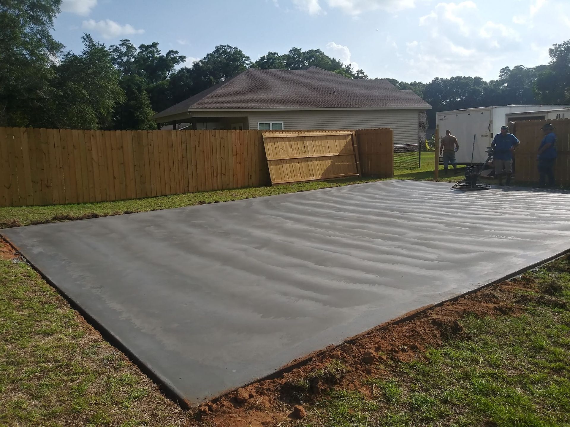 Large freshly poured concrete slab in residential backyard, smooth troweled surface with visible finishing lines, bordered by grass and soil, workers operating power trowel near wooden privacy fence.