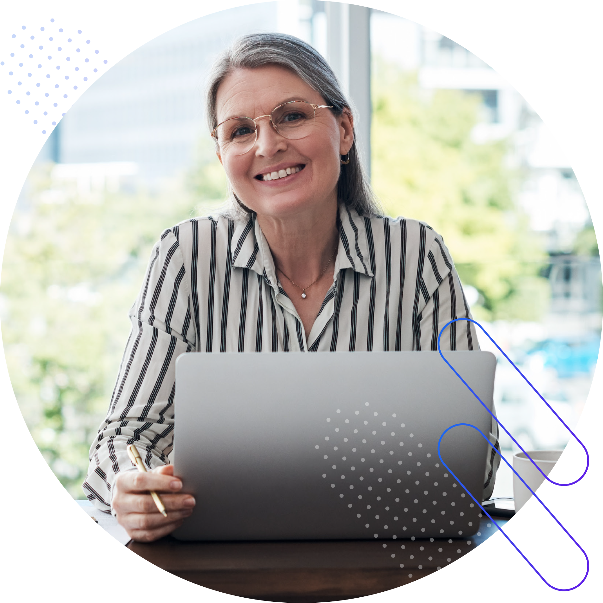 A middle-aged woman with gray hair and glasses smiles at the camera while sitting at a table with a laptop in front of her. She is wearing a black and white striped shirt and a gold necklace. Her right hand is holding a gold pen over the laptop. The background is blurred, suggesting an office or cafe setting with natural light coming from a window.