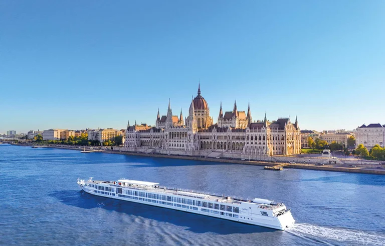 A large white river cruise ship sails on a blue river in front of the Hungarian Parliament Building in Budapest under a clear blue sky.