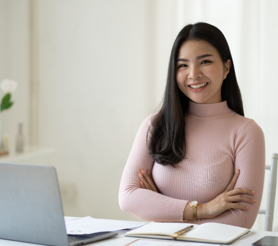 A young Asian woman is smiling and looking at the camera with her arms crossed. She is wearing a pin 