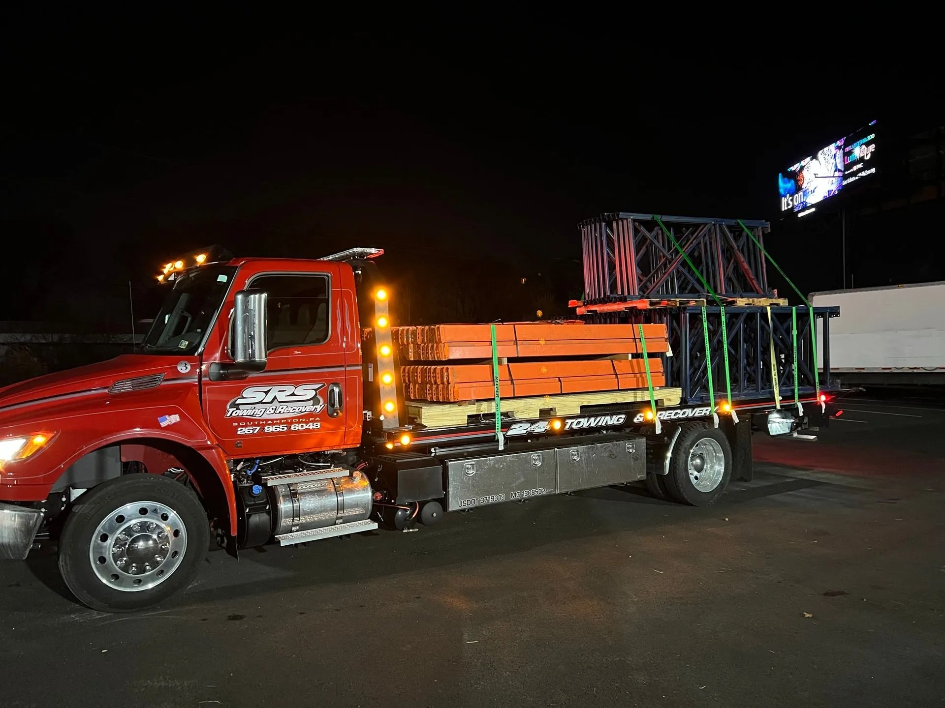A red SRS Towing & Recovery flatbed truck parked at night, carrying industrial metal racks and orange beams, tightly secured with green straps. The truck's amber lights are on, illuminating the dark surroundings.