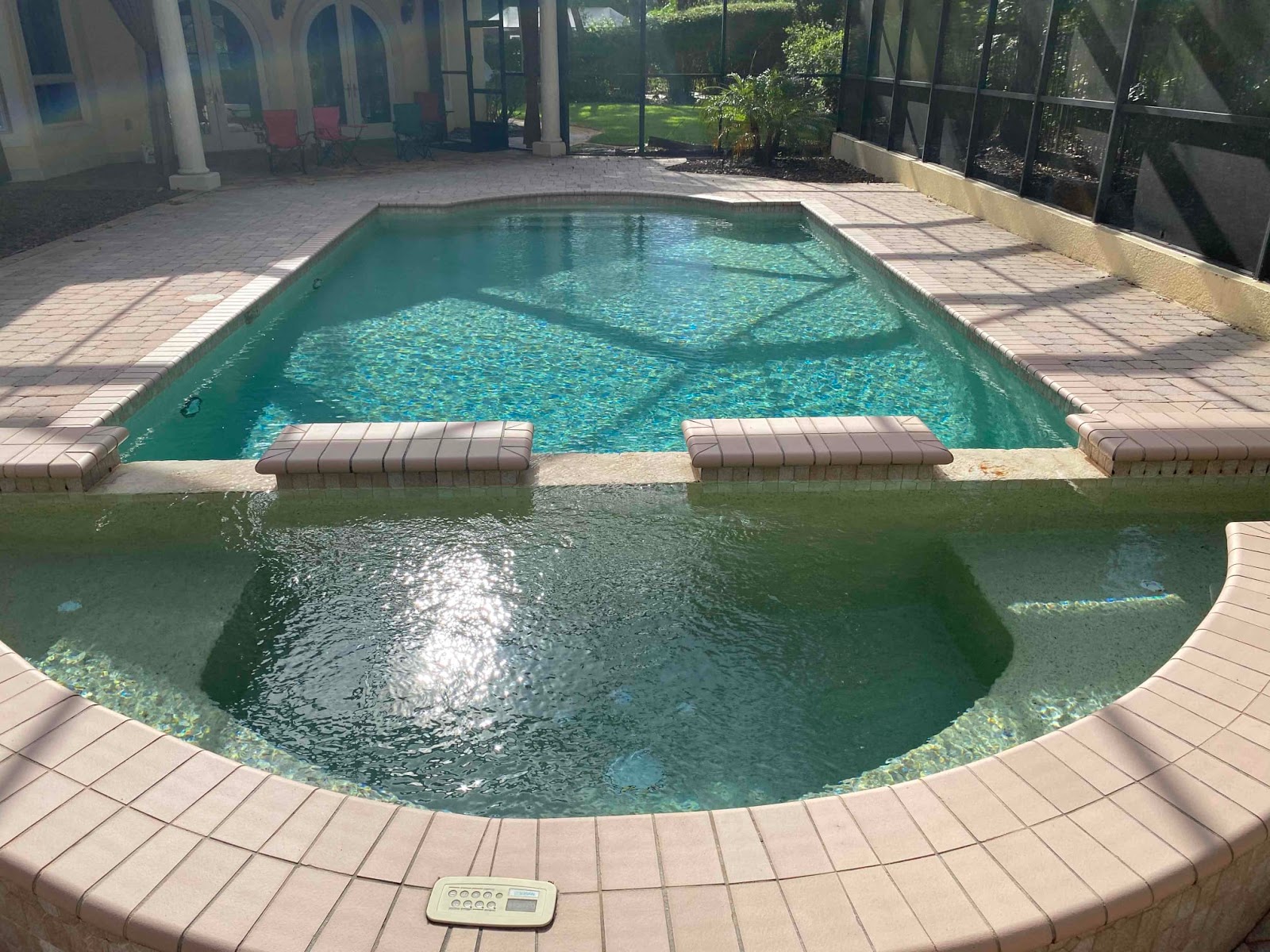 Screen-enclosed backyard swimming pool with an attached raised spa, green-blue water, light paver decking, and a private outdoor setting.