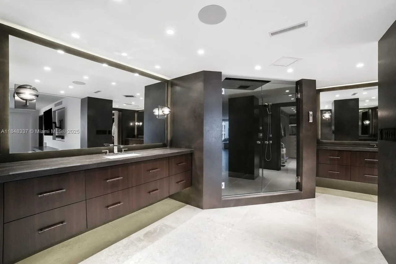 A spacious and modern master bathroom featuring a large vanity with a sink and dark wood cabinetry, a glass-enclosed shower with modern fixtures, and a large wall mirror reflecting the room.