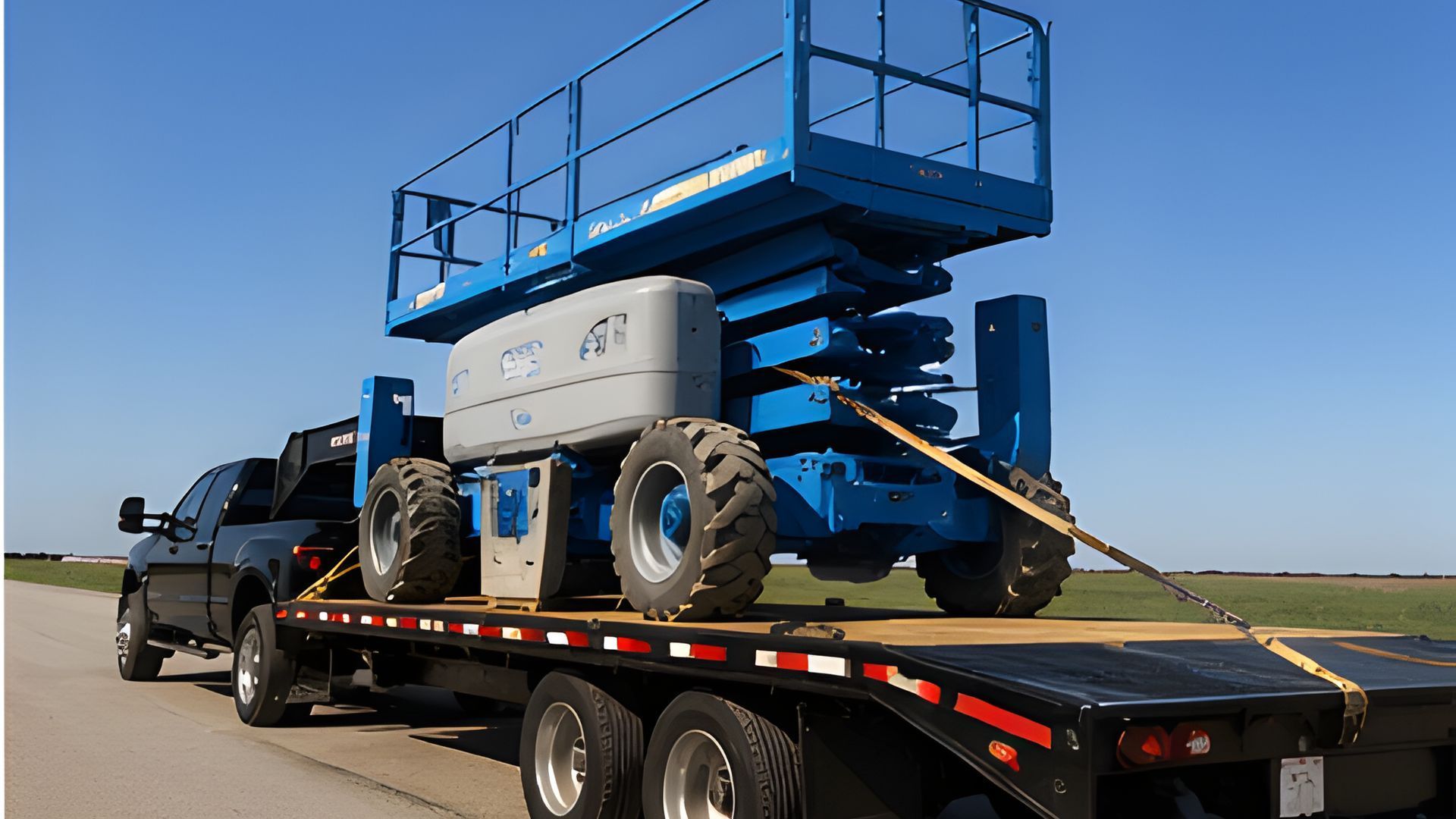 Blue scissor lift secured on a flatbed trailer for transport. Equipment hauling service moving aerial work platforms safely between construction sites and industrial project locations.