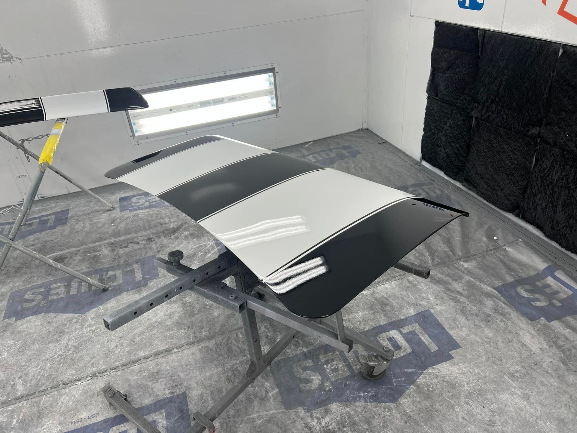 A detached body panel displays a glossy black finish accented with white racing stripes. The piece is drying after professional painting and custom automotive restoration services.