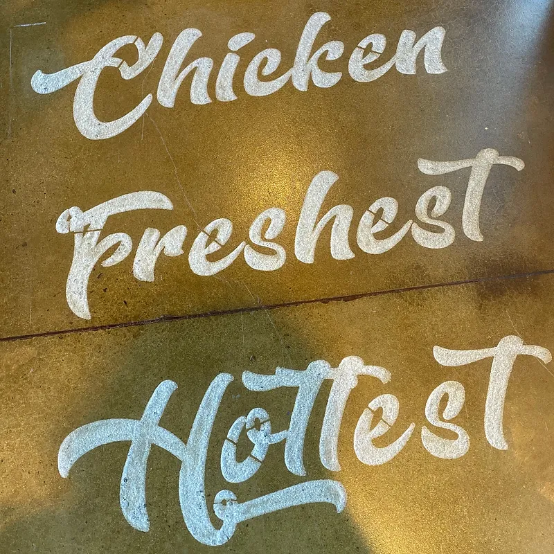 White text reads "Chicken", "Freshest", and "Hottest" stenciled onto a textured, metallic-gold surface. The text is in a flowing, script font with some stencil breaks in the letters.
