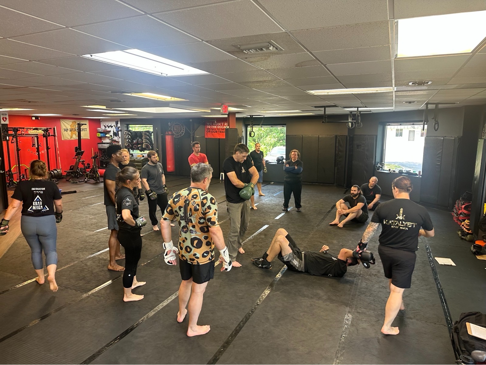 An adult Krav Maga class on the mats at Katalyst Kombatives in Virginia Beach while an instructor demonstrates a ground defense.