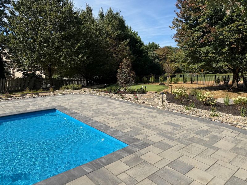 Modern backyard featuring a paved patio surrounding a swimming pool with clean stonework and landscaped borders. The design includes trees, plants, and organized layout, showcasing professional hardscaping and outdoor living space installation for residential property enhancement.