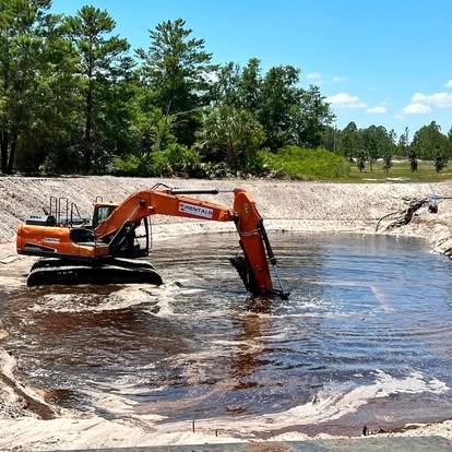 An orange excavator positioned inside a deep sandy basin dredging dark water.