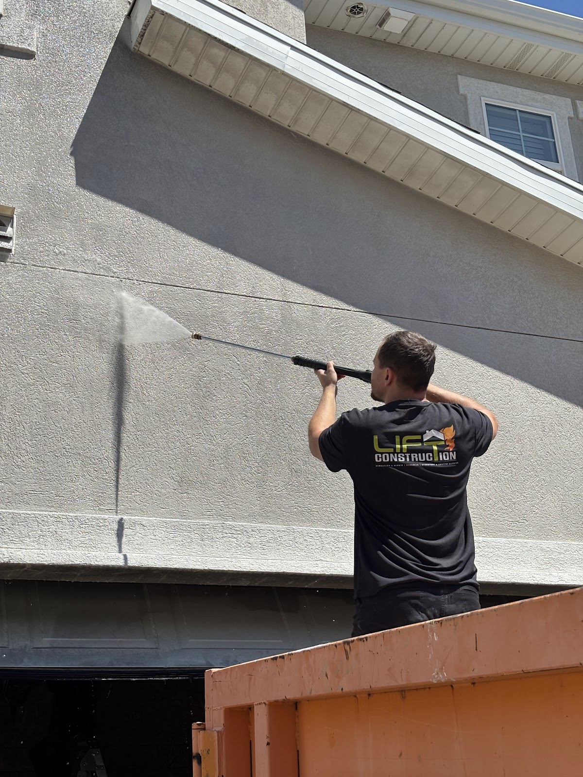A man in a black t-shirt with a company logo on the back is pressure washing the side of a building. 
