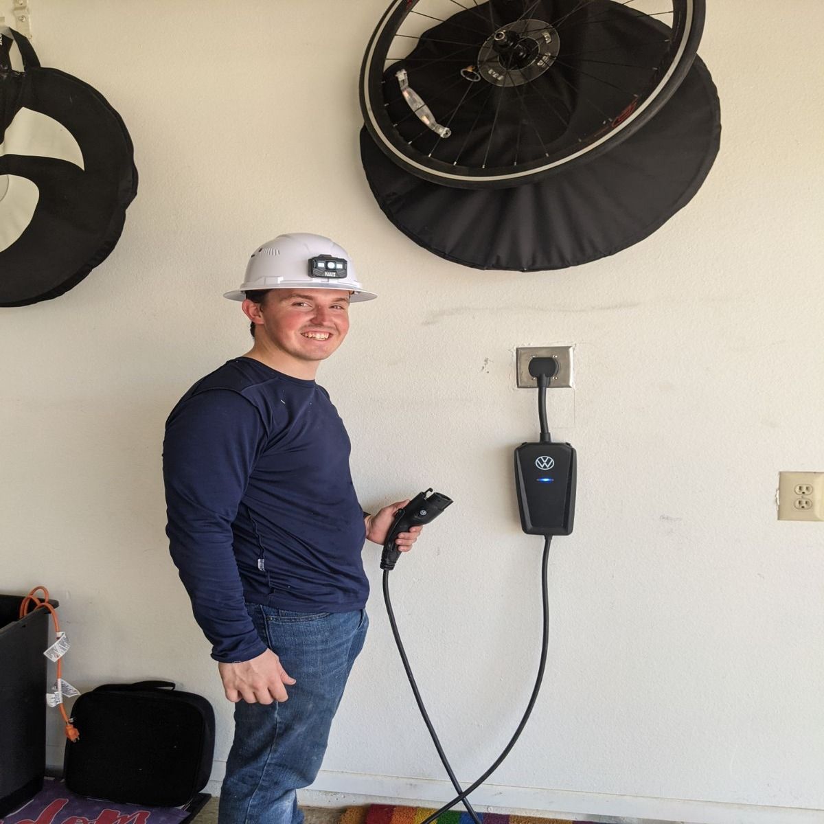 An electrician stands beside a wall-mounted Volkswagen electric vehicle charger in a residential garage, holding the charging connector while posing near bicycles and equipment after completing the EV installation work.
