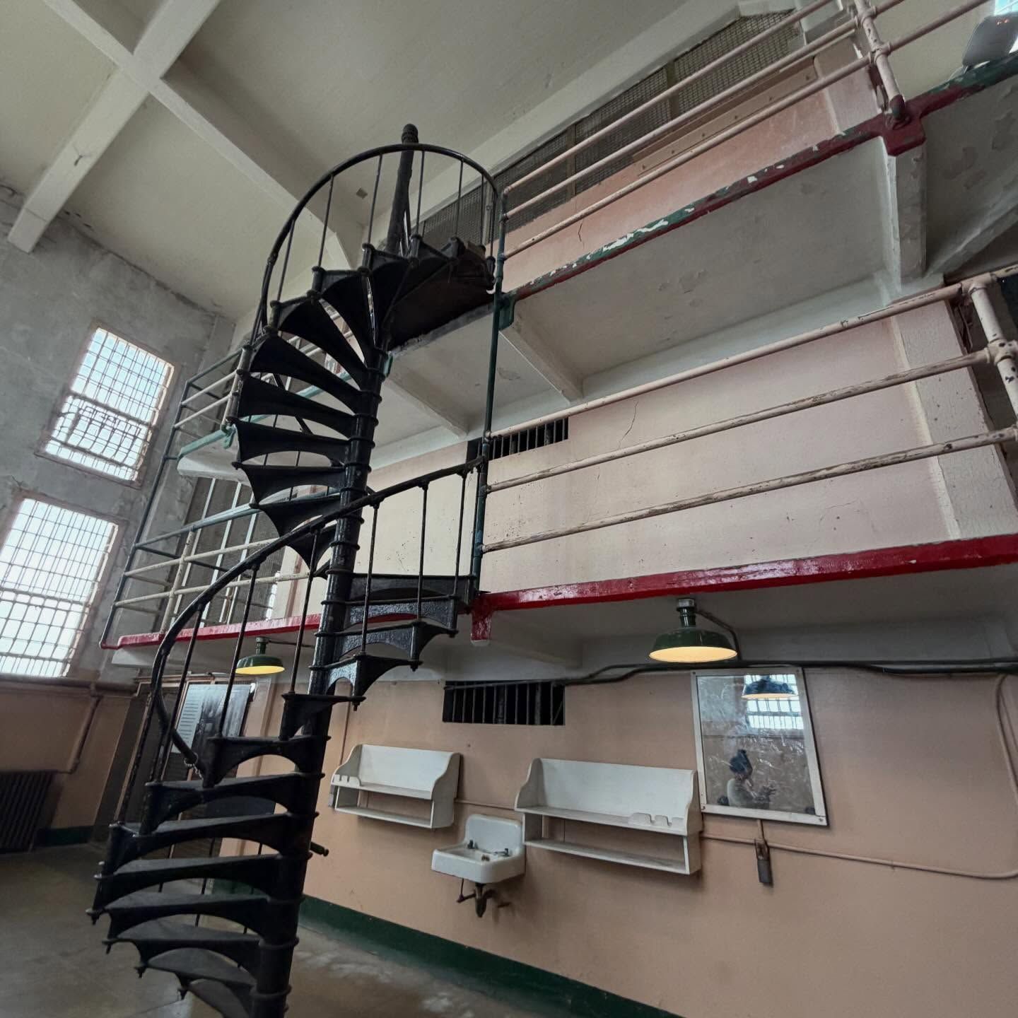Interior of a prison cell featuring a spiral metal staircase leading to an upper level, with barred windows, industrial lighting, wall-mounted sink and shelves, showcasing confined living space and institutional design.