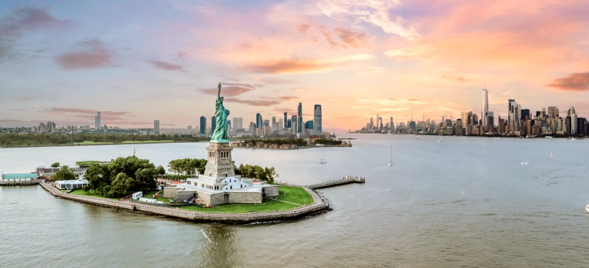 The Statue of Liberty in the foreground with New York Harbour and the city skyline in the background at sunset.