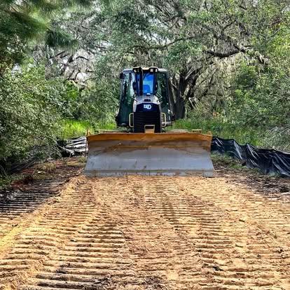 A CAT bulldozer driving forward on a cleared sandy path through a wooded area lined with black silt fencing.