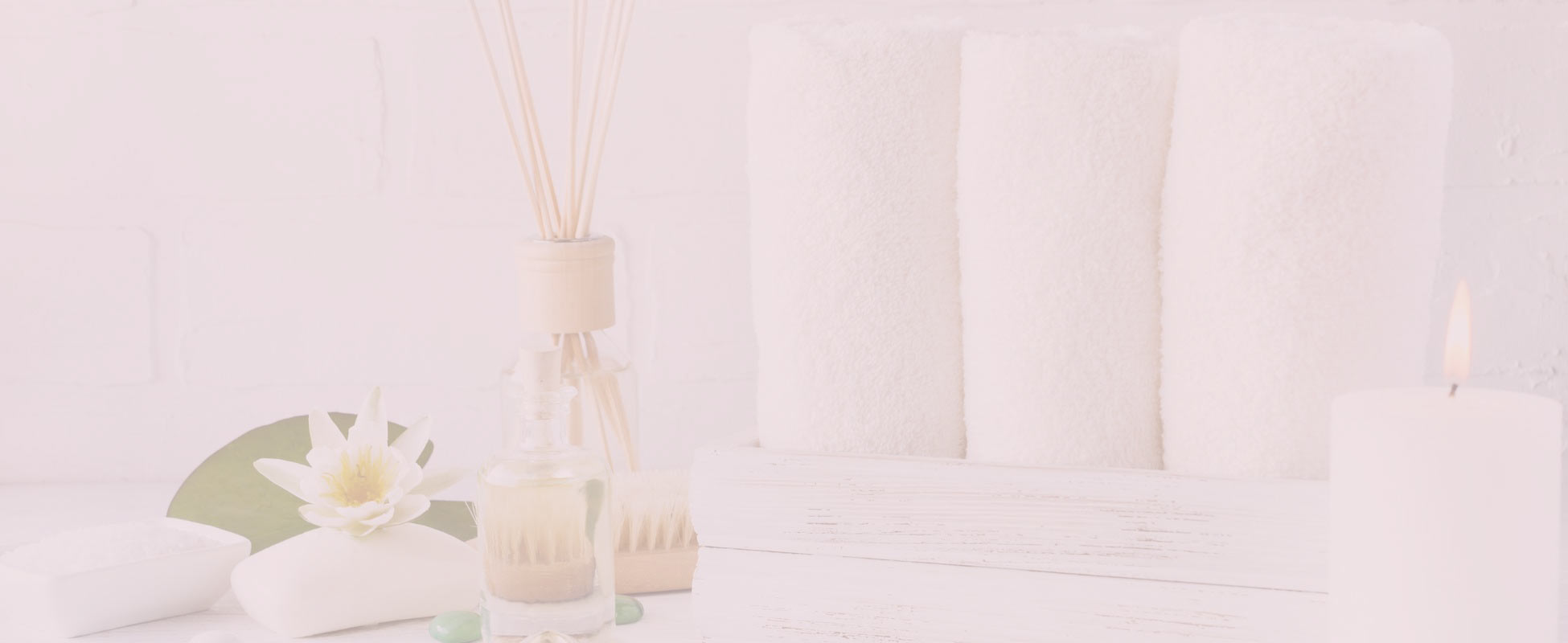 A still life arrangement for a spa or bath setting. The scene features a lit white pillar candle on the right, and a row of neatly folded white towels in the background. To the left, there is a glass bottle of diffuser oil with wooden reeds, a white bar of soap, a white dish filled with bath salts, a single white water lily on a green lily pad, and a small wooden brush. The overall aesthetic is clean, calming, and serene.
