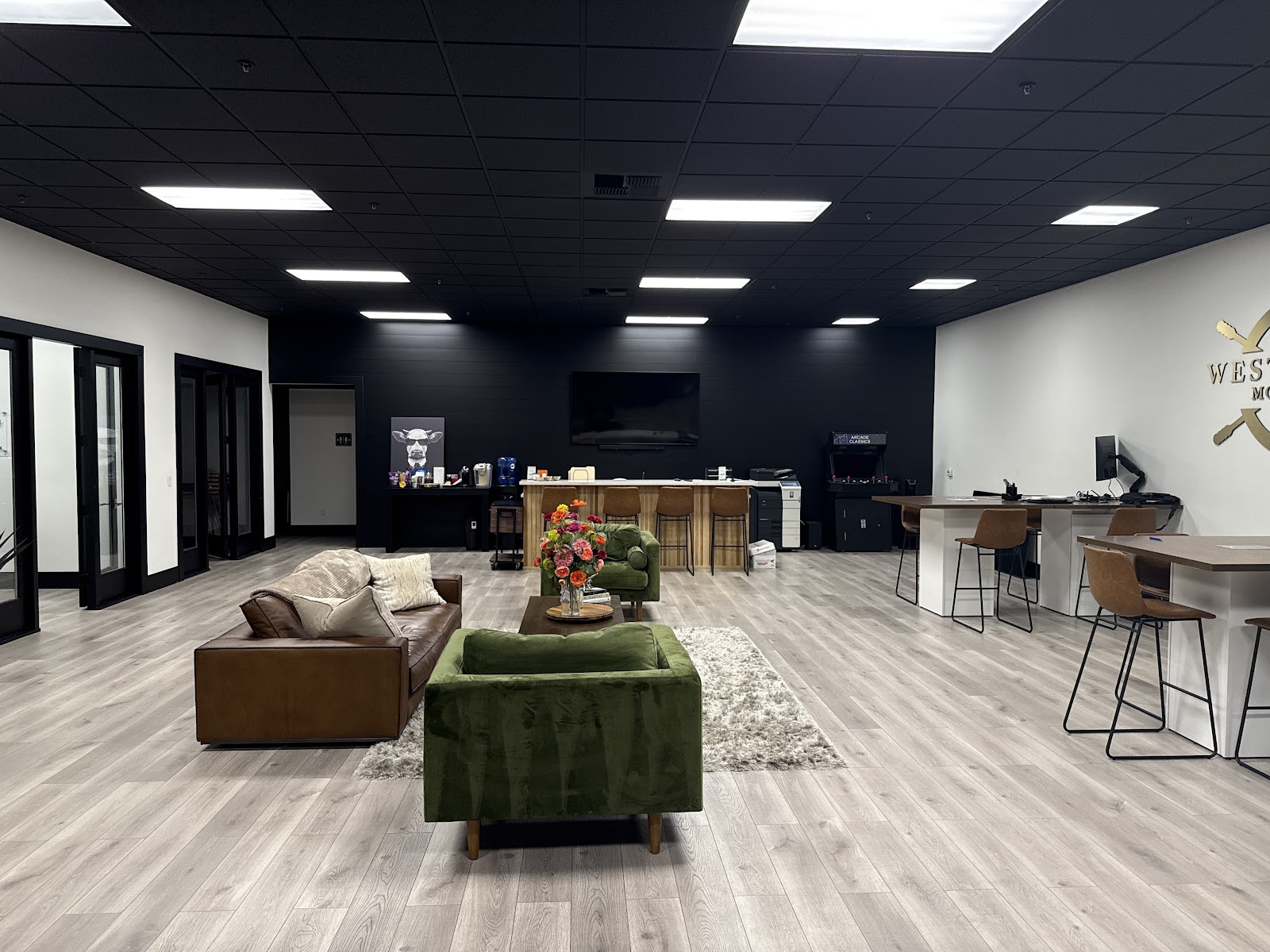An office waiting area with a lounge area featuring a brown leather sofa and a green armchair, a coffee table with a vase of flowers, and a textured rug. Beyond the lounge area, there's a bar with stools, a large TV mounted on a black wall, and desks with computers and chairs. A vintage arcade machine and a printer are also visible. The walls are white and black, and the ceiling is black with fluorescent lighting. A logo is mounted on the white wall.