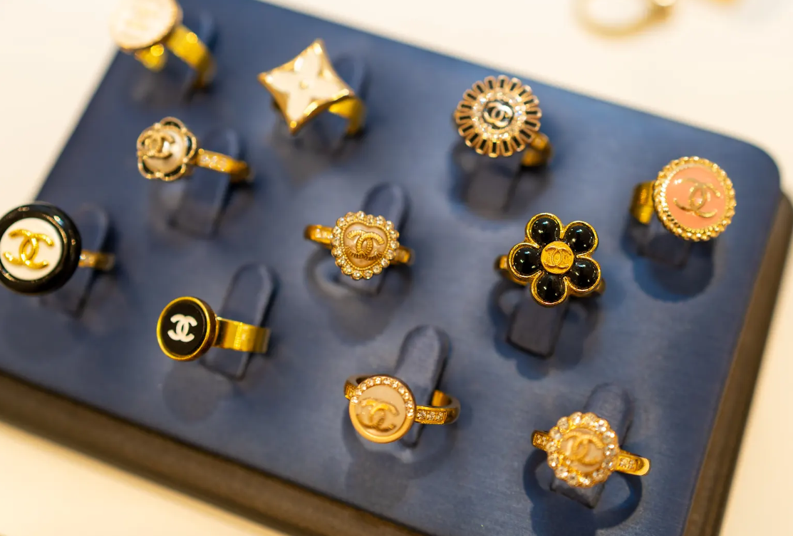 Gold-tone statement rings with floral shapes, logo details, and sparkling accents shine on a display at Eliana Curated. Explore distinctive jewelry styles in Old Town Alexandria, Virginia. Shop local.