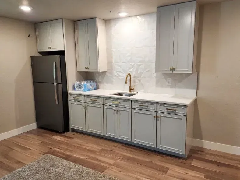 Beautiful basement kitchenette with gray shaker cabinets, quartz countertops, decorative backsplash, modern faucet, stainless refrigerator, and luxury vinyl flooring, providing convenience and functionality for homeowners.