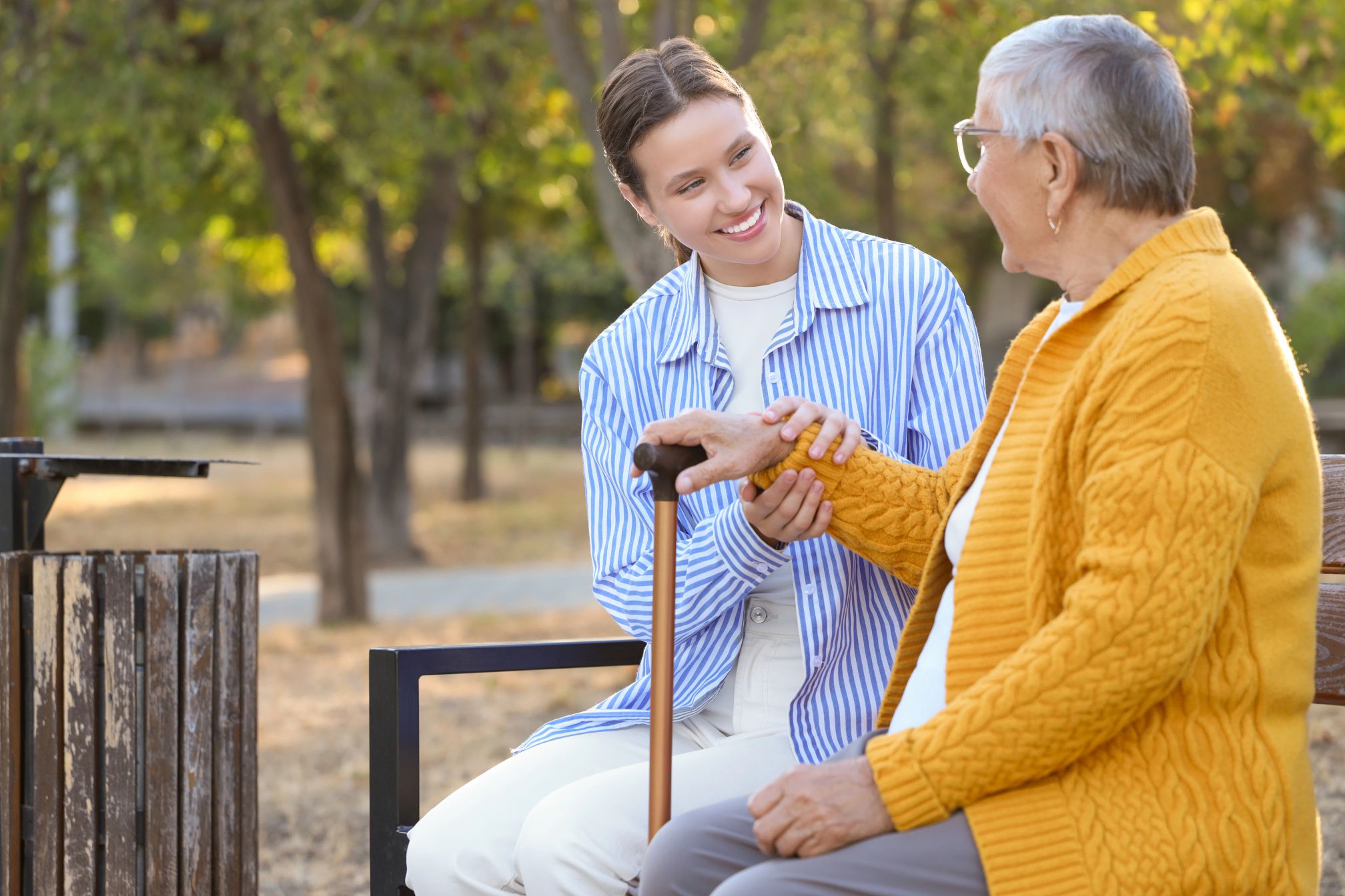 A young woman in a blue and white striped shirt and a senior woman in a yellow cardigan sit on a park bench and talk. The younger woman is holding the older woman's hand and the older woman is holding a cane. Trees and a trash can are visible in the background.