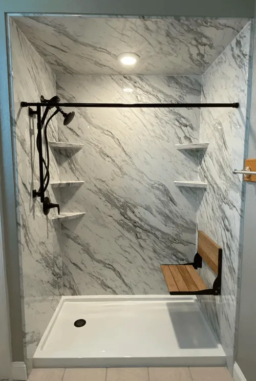 A clean, modern, and accessible shower stall featuring marble-patterned walls, a white shower base, a fold-down wooden seat, and a multi-head shower system. The stall is well-lit by recessed ceiling lights and appears to be recently renovated.