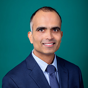 Male doctor in white medical coat smiling in front of teal blue backdrop. Rajesh Srinivasan, MD Springfield (447)448-3041