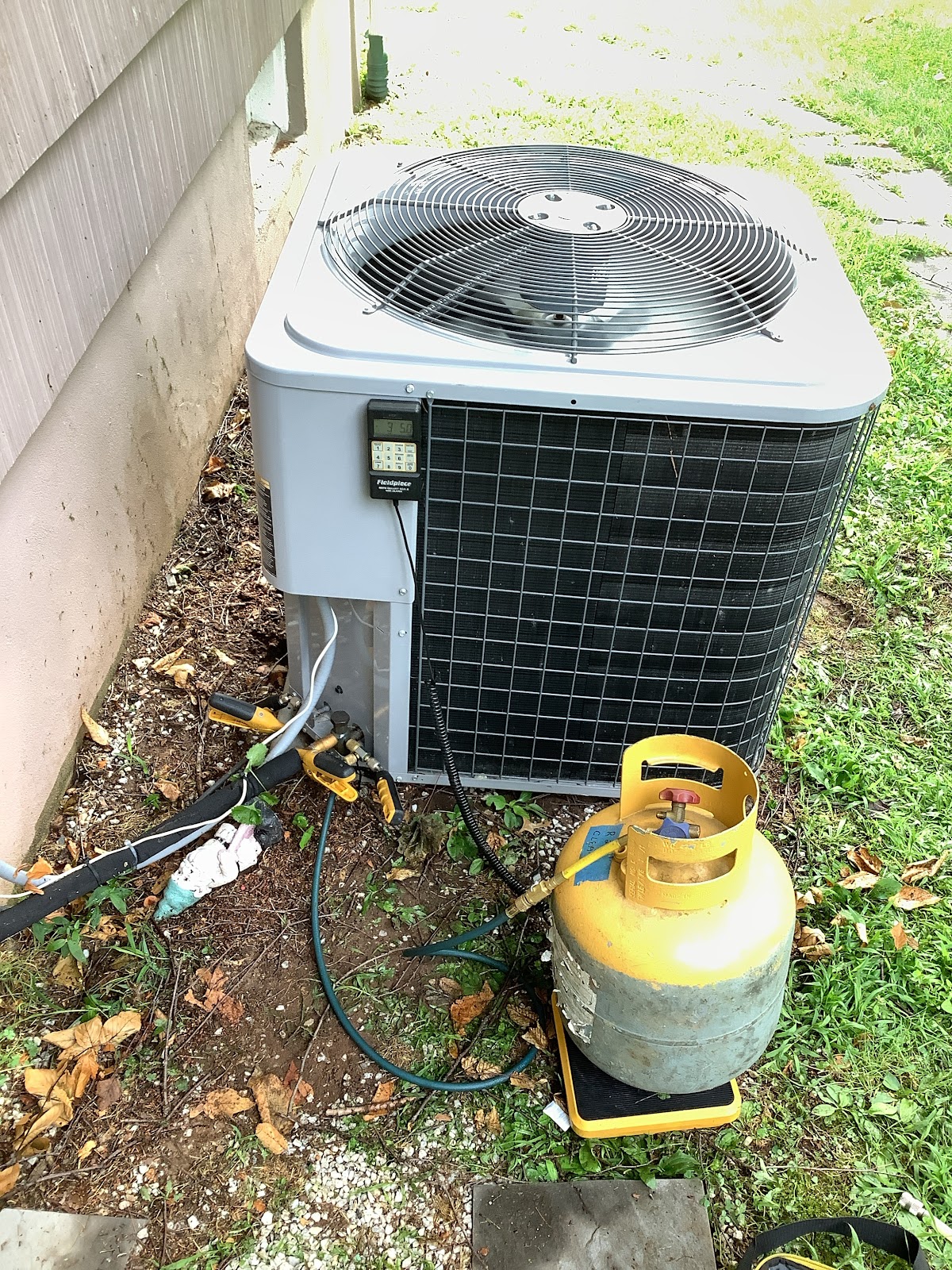 An outdoor air conditioning unit is being serviced with a yellow refrigerant tank and gauges attached. The unit is a light gray color with a large fan grille on top and a black metal grille on the side. A digital display, possibly a manifold gauge set, is attached to the side of the unit. A yellow propane-like tank is placed on a yellow scale next to the AC unit, connected by hoses. The surrounding area is covered in grass, dirt, and fallen leaves, with the side of a building visible on the left.