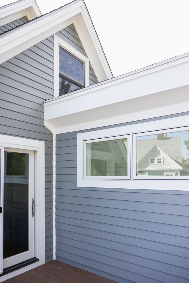 A close-up view of the exterior of a modern house with gray horizontal siding and white trim around windows and rooflines. A sliding glass door is visible on the left, and a set of two horizontal windows is on the right. The windows reflect the roof of a house across the street. The sky is overcast and bright white.