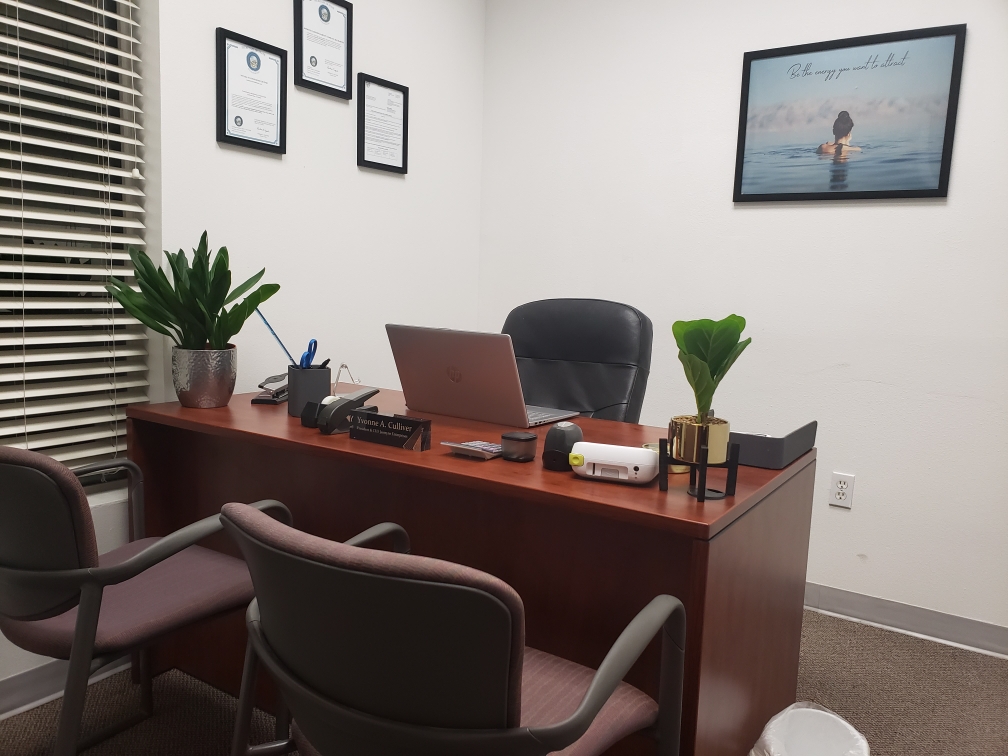 A clean and organized office desk with a laptop, plants, and framed documents on the wall. The desk  