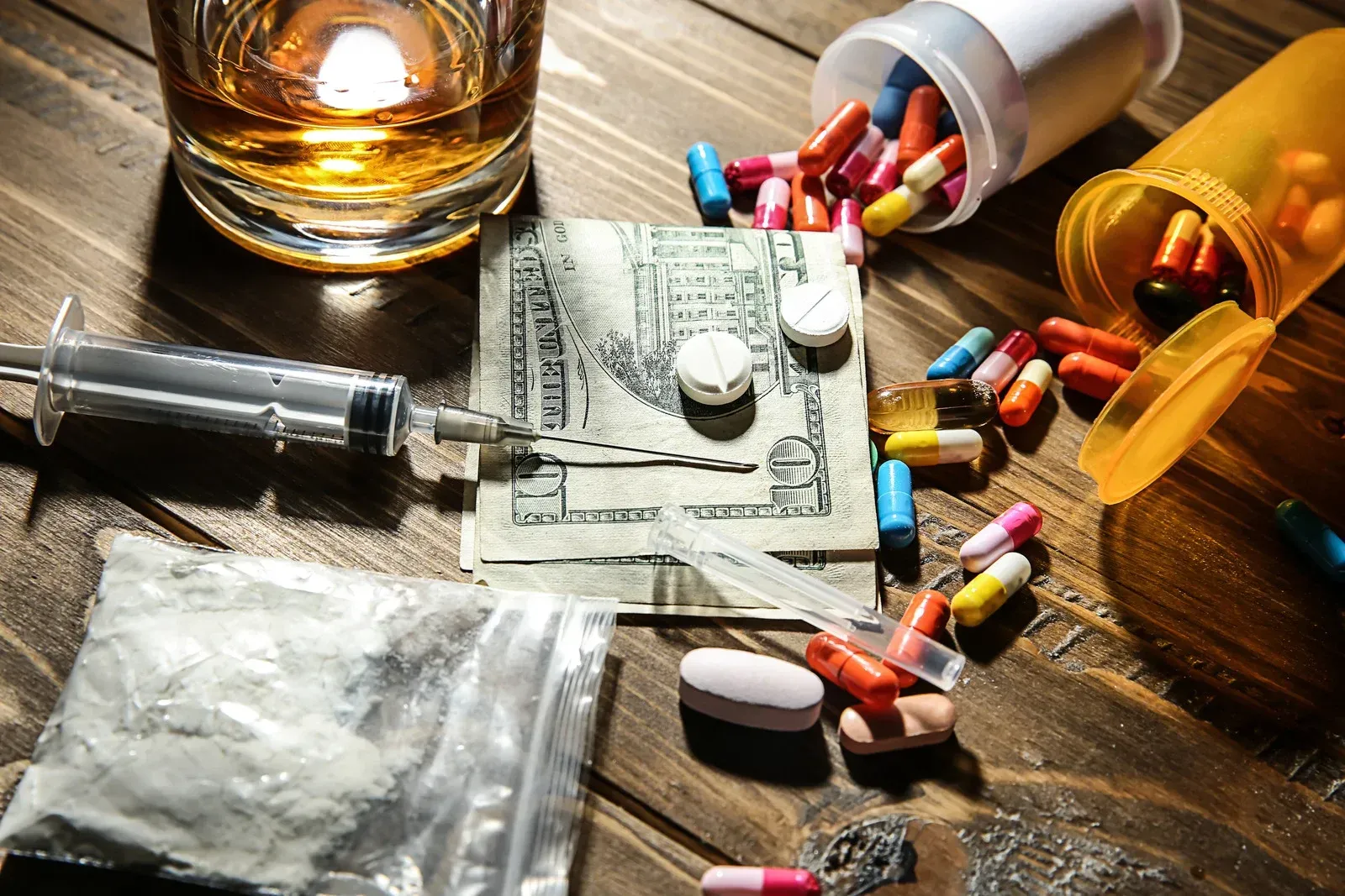The image displays a collection of drug paraphernalia and substances on a wooden surface. This includes various pills and capsules scattered near overturned prescription bottles, a syringe, a small bag of white powder, and folded US dollar bills next to white tablets. A glass of amber liquid is also present.