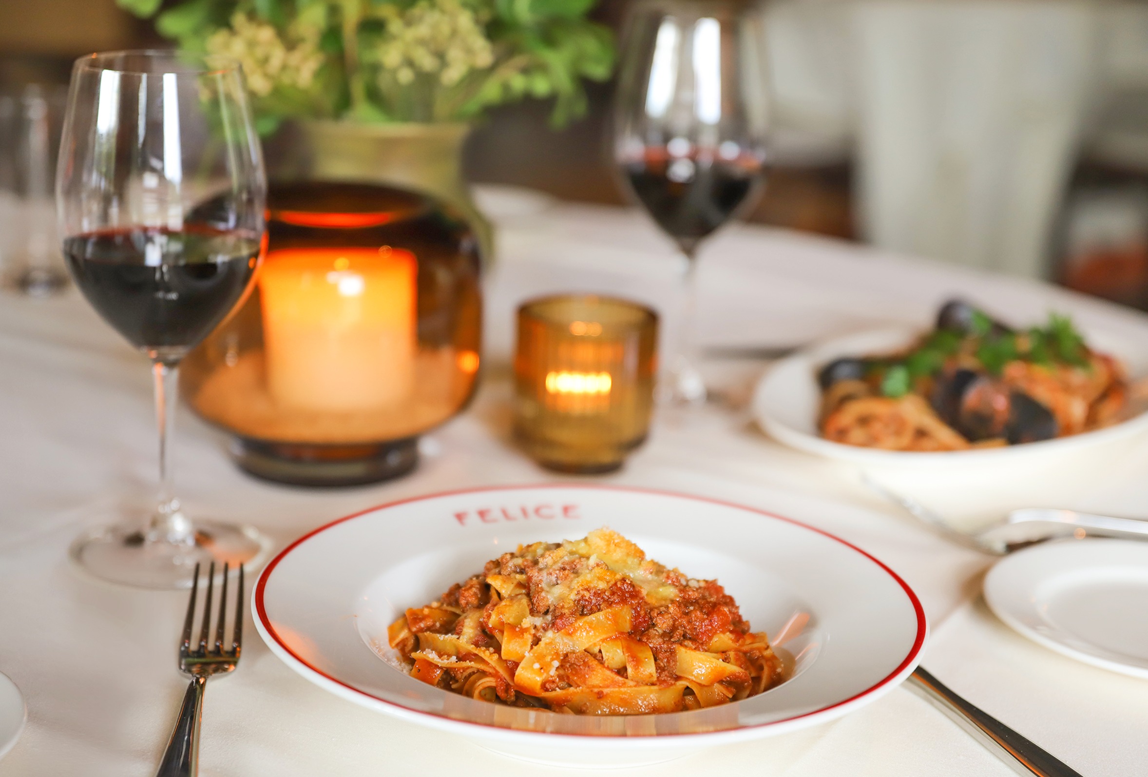 The warm, intimate atmosphere of a dinner at Felice, showing a detailed shot of authentic meat pasta and mussels in the background, illuminated by soft amber candlelight.