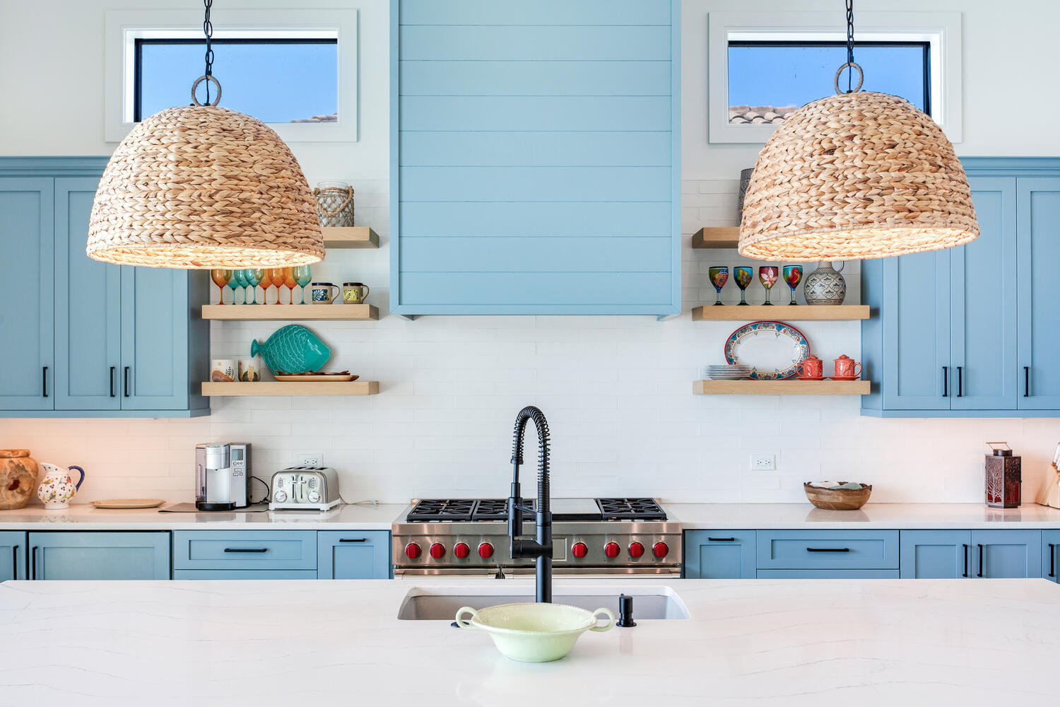 Bright coastal kitchen detail with light blue cabinetry, woven pendant lights, white countertops, and a stylish sink area with open shelving.