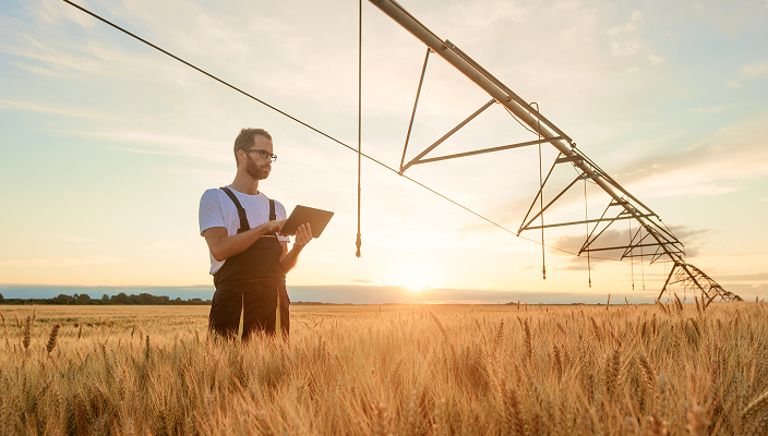 Man in overalls surveying a field with an industrial element overhead.