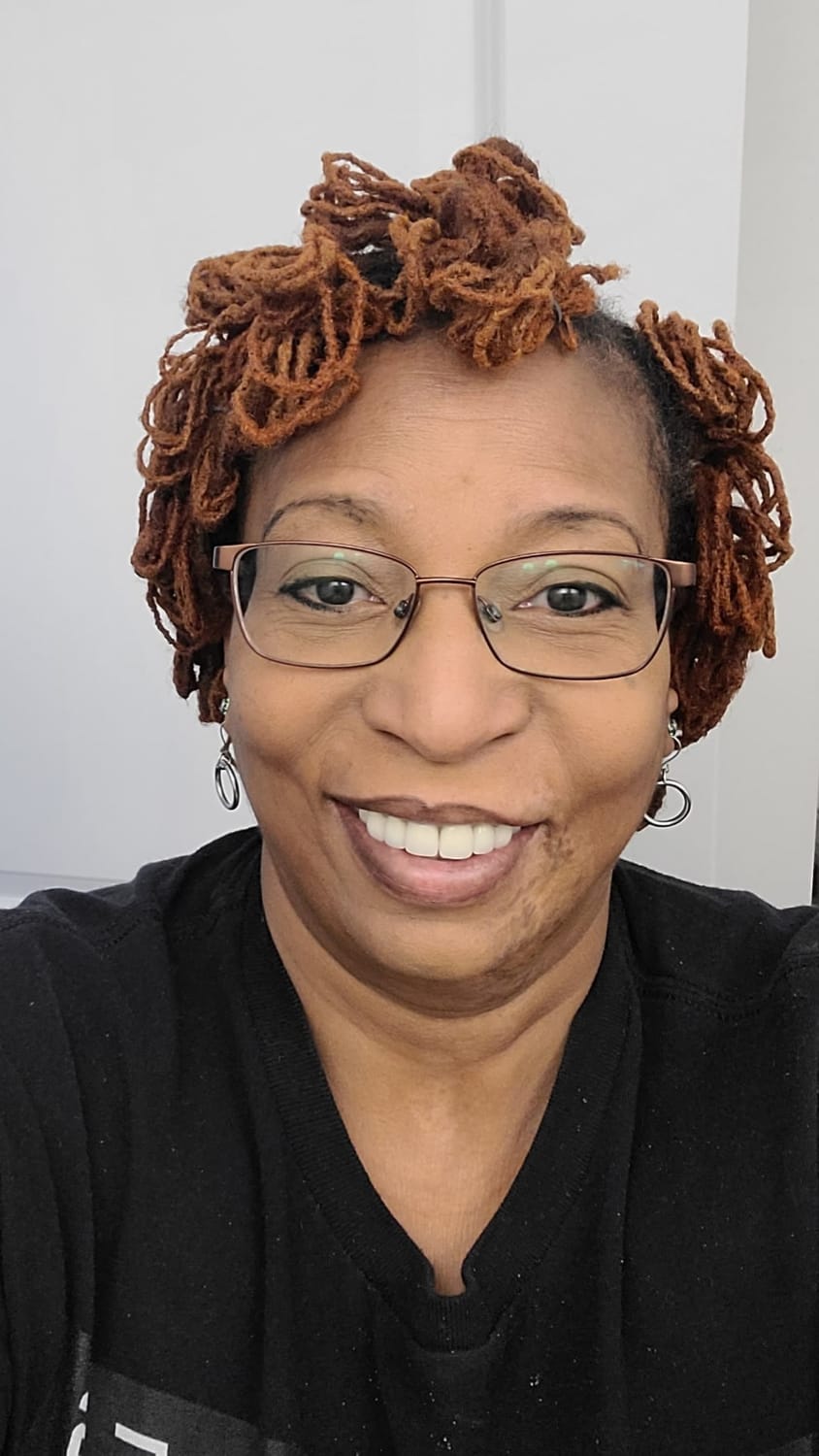 A close-up, waist-up photo of Tracy Denise Anderson smiling and wearing glasses. Her hair is styled in short, reddish-brown locs. She is wearing a black t-shirt and silver hoop earrings. The background is a plain white wall.