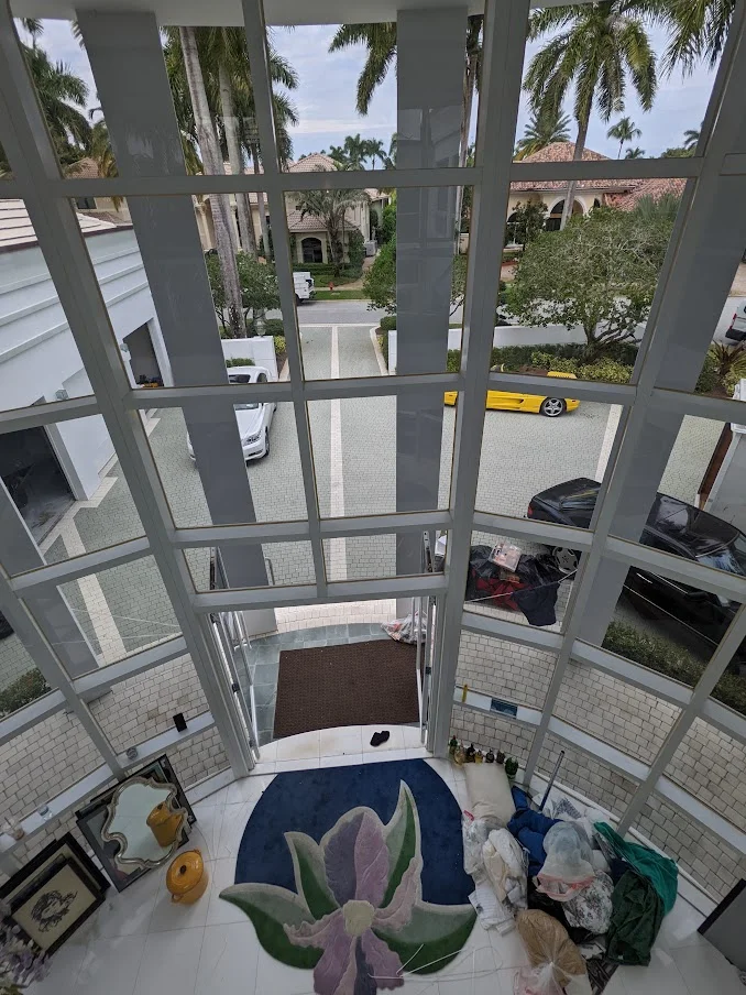 A high-angle view from inside a modern house with large windows looking out onto a driveway and several houses. Two cars are visible on the driveway: a silver sedan and a yellow sports car. In the foreground, a large rug with a floral design is on the floor, along with several items that appear to be moving boxes or stored items. The architecture features a lot of glass and clean lines.